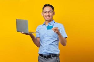 leende ung asiatisk man asiatisk i glasögon håller laptop och visar kreditkort isolerad på gul bakgrund. affärsman och entreprenör koncept foto