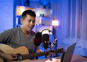 asiatisk man youtuber live streaming prestanda spelar gitarr och sjunger en låt. asiatisk man som undervisar i gitarr och sjunger online. musiker som spelar in musik med laptop och spelar akustisk gitarr. foto