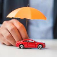 affärsman hand håller paraply och täcka röd bil leksak på bordet. bilförsäkring, garanti, reparation, finansiell, bank och pengar koncept foto
