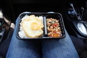ångat ris med stekt ägg och kycklingbasilika i en plastförpackning placerad på en kvinnas ben i bilen. foto