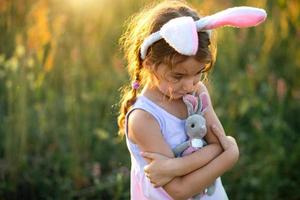 söt 5-årig tjej med kaninöron kramar försiktigt en leksakskanin i naturen i ett blommande fält på sommaren med gyllene solljus. påsk, påskhare, barndom, lyckligt barn, våren. foto