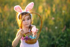 söt rolig tjej med målade påskägg på våren i naturen i ett fält med gyllene solljus och blommor. påskhelg, påskharen med öron, färgglada ägg i en korg. livsstil foto