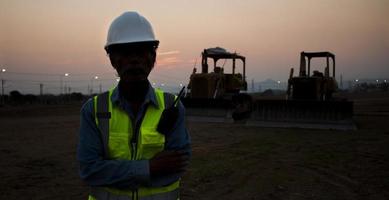 siluett senior ingenjör bär hjälm och reflexväst på solnedgången bakgrund och larvband bulldozer. foto