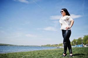 brunett flicka på kvinnors läderbyxor och vit blus, solglasögon, poserade på geen gräs mot stranden av sjön. foto