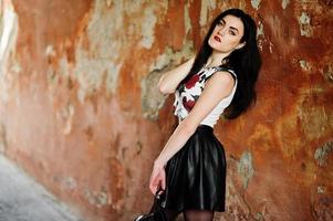 ung goth flicka på svart läder kjol med ryggsäck poserade mot grunge vägg. foto