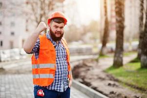 porträtt av arg skägg arbetare man kostym byggnadsarbetare i säkerhet orange hjälm mot trottoar med visar armar. foto