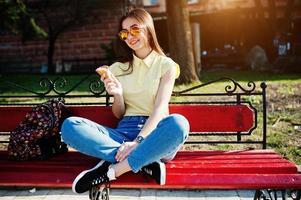 ung tonårsflicka sitter på bänken med glass vid händerna, bär på gul t-shirt, jeans och solglasögon. foto