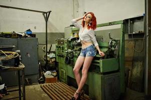 rödhårig flicka på korta jeansshorts och vit blus poserade vid industriell maskin på fabriken. foto