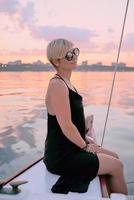 vacker blond glad kvinna i solglasögon och lång svart klänning njuter av solnedgången på yachten i havet. resor, äventyr, chill, avkoppling koncept foto
