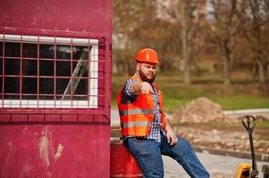 arg skägg arbetare man kostym byggnadsarbetare i säkerhet orange hjälm sitter på röda fat och visar finger att arbeta. foto