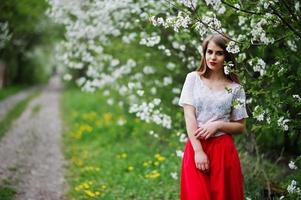 porträtt av vacker flicka med röda läppar på våren blossom trädgård, bära på röd klänning och vit blus. foto