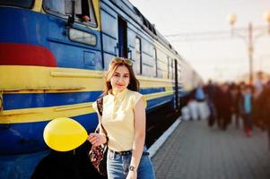 ung tonårsflicka som står på perrongen vid tågstationen med ballong till hands, bär på gul t-shirt, jeans och solglasögon, med ryggsäck. foto