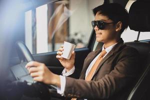 stilig asiatisk affärsman i solglasögon kör bil medan han håller en kopp kaffe foto