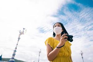 vuxen asiatisk kvinna bär skyddande ansiktsmask och gul klänning håller kamera för att ta foto
