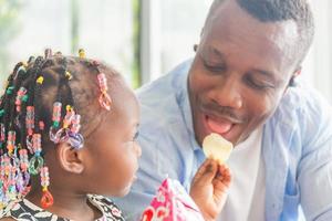 söt liten flicka tittar på hennes pappa som äter mellanmål, glada afroamerikanska far och dotter som leker i vardagsrummet, familjekoncept för lycka foto