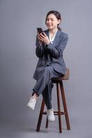 ung asiatisk affärskvinna sitter på stol och använder smartphone på grå bakgrund foto