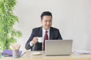 chefen för en asiatisk man som smuttar på kaffe på sitt skrivbord på ett strålande sätt. foto
