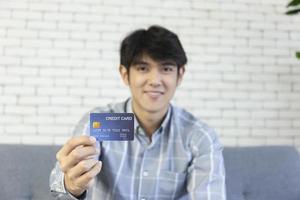 en ung asiatisk man som sitter på en soffa med en hand som håller ett kreditkort foto