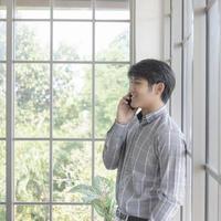 en ung asiatisk affärsman står på balkongen i ett fönster och pratar i telefon. foto