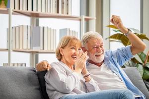porträtt av lyckliga äldre par hejar på sitt favoritlag medan de tittar på fotbollsmatch i vardagsrummet, äldre kvinna och en man som kopplar av på mysig soffa hemma, lyckliga familjekoncept foto