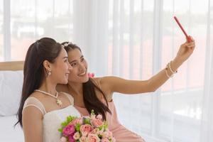 brudens vän använder en smartphone för att ta en selfie av en asiatisk brud som håller i en bukett vackra blommor i en bröllopsklänning i omklädningsrummet på sängen.