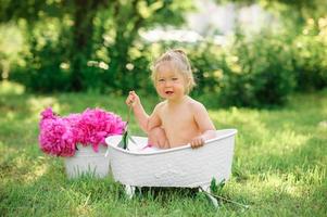 glad toddler flicka tar ett mjölk bad med kronblad. liten flicka i ett mjölkbad på en grön bakgrund. buketter av rosa pioner. babybadning. hygien och omsorg för små barn. foto