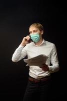 en ung man i medicinsk mask grälar i telefonen och analyserar de mottagna dokumenten. konceptet att hitta en lösning på affärsproblem på grund av epidemin av coranavirus foto