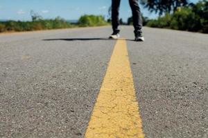 man fötter stående på asfaltvägen med gula markeringslinjer. man bär sneakers och jeans. foto