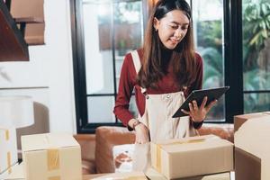 sme småföretag asiatisk kvinnlig entreprenör eller butiksägare kontrollera kundorder på digital surfplatta medan du packar lådor hemma. använder social för marknadsföring online shopping, e-handelskoncept foto