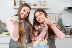 glad mor och dotter visar leende och visar hemlagad tårta eller cupcakes tittar på kameran i köket. familjeaktivitet bakning bageri hemma på helgen foto