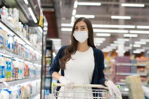 kvinna som bär medicinsk ansiktsmask och gummihandske push kundvagn i stormarknad varuhus. flicka tittar på livsmedelsbutik hyllan. ny normal livsstil under coronavirus eller covid-pandemi foto