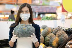 kvinna som bär medicinsk ansiktsmask och gummihandske håller pumpa förbereder sig för halloween-semester på grönsaksvaruhuset. shopping i snabbköpet i en ny normal livsstil foto