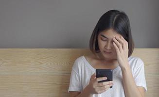 upprörd ung kvinna som sitter på sängen, håller smartphone i händerna, fick meddelande med dåliga nyheter. foto