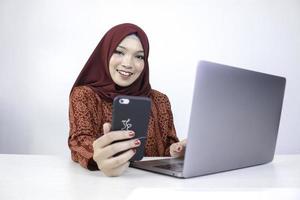 ung asiatisk islam kvinna som bär huvudduk ler på en mobiltelefon med laptop på bordet. foto