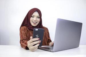 ung asiatisk islam kvinna som bär huvudduk ler på en mobiltelefon med laptop på bordet. foto
