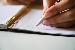 mans hand skriven i en spiral anteckningsblock placerad på ett träbord med föremål. man hand med penna skriver på anteckningsboken foto