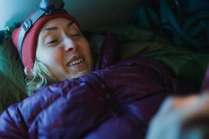 en kvinna med pannlampa ligger i en sovsäck i ett tält och håller i en smartphone foto