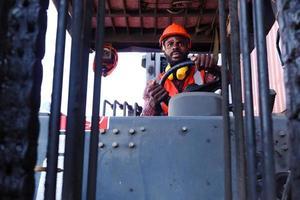 industriarbetare man bär säkerhet ljus neonröd väst och hjälm kör gaffeltruck bil på fabriken fabrik industri, afrikansk amerikansk ingenjör manlig arbetar på logistisk frakt last container gård. foto