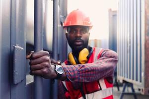 porträtt av afroamerikansk ung ingenjör arbetare man bär säkerhets ljus neon röd färg väst och hjälm, försöker öppna en container dörr på logistisk last container varv. foto