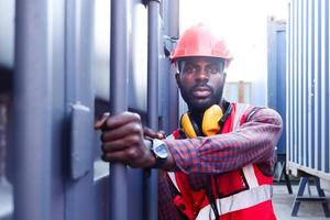 porträtt av afroamerikansk ung ingenjör arbetare man bär säkerhets ljus neon röd färg väst och hjälm, försöker öppna en container dörr på logistisk last container varv. foto