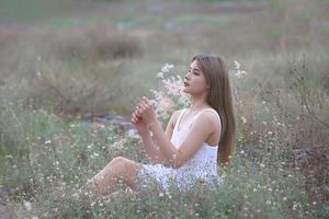 vacker ung kvinna sitter på fältet i grönt gräs och blåser maskros. utomhus. njut av naturen. frisk leende flicka på våren gräsmatta. allergifritt koncept. frihet foto