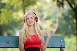 vacker blond kvinna står medan du lyssnar på musik i hörlurar i parken. foto