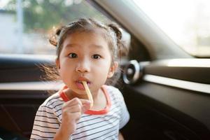 liten asiatisk ung flicka som stannar i bilen och äter stekt potatischips, härlig flicka njuter av att äta mellanmål när hon reser med bilen. foto