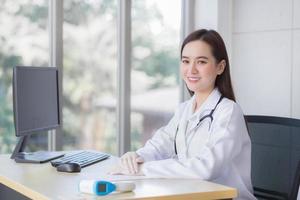 professionella asiatiska vackra unga leende kvinnliga läkare bär gummihandske sitter och tittar på kameran medan du väntar på patientundersökning på kontoret på sjukhuset. foto