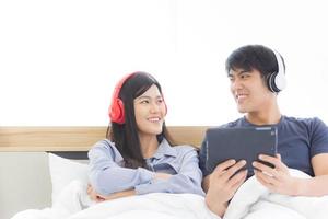 ett lyckligt ungt asiatiskt par sitter i sängen och njuter av att lyssna på låtar eller titta på film på en surfplatta på en vit bakgrund. foto