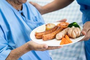 asiatisk senior eller äldre gammal dam kvinna patienten äter frukost hälsosam mat med hopp och glad medan du sitter och hungrig på sängen på sjukhuset. foto