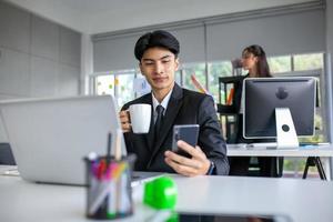 porträtt av asiatisk stilig man som arbetar på kontoret, med smartphone och dator på bordet. foto