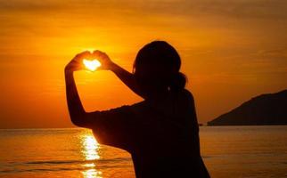 siluett av en flicka som räcker upp handen för att få ett hjärta att sätta på solen. vid havet. morgonsol. på havet. ljusa toner av orange. foto