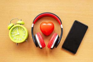 hörlurar och väckarklocka och smartphone och rött hjärta på trä skrivbord. musikaliskt koncept foto