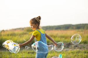 en flicka i en jeansoverall blåser såpbubblor på sommaren i ett fält vid solnedgången. internationella barndagen, glada barn, utomhusaktiviteter. sommar bakgrund. hälsosam och miljövänlig livsstil foto